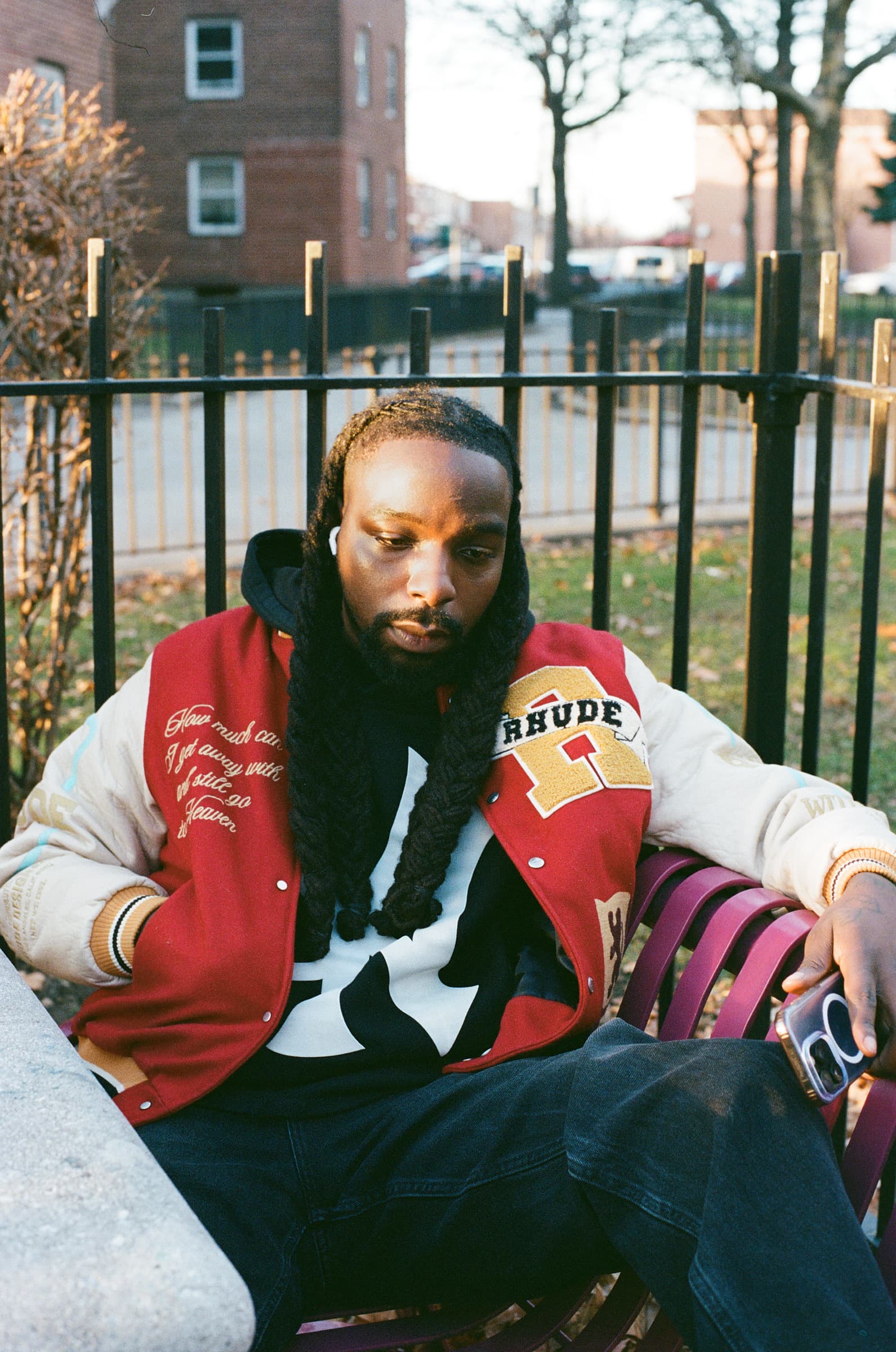Portrait of a man in a varsity jacket and black hoodie, sitting on a park bench in front of NYCHA housing