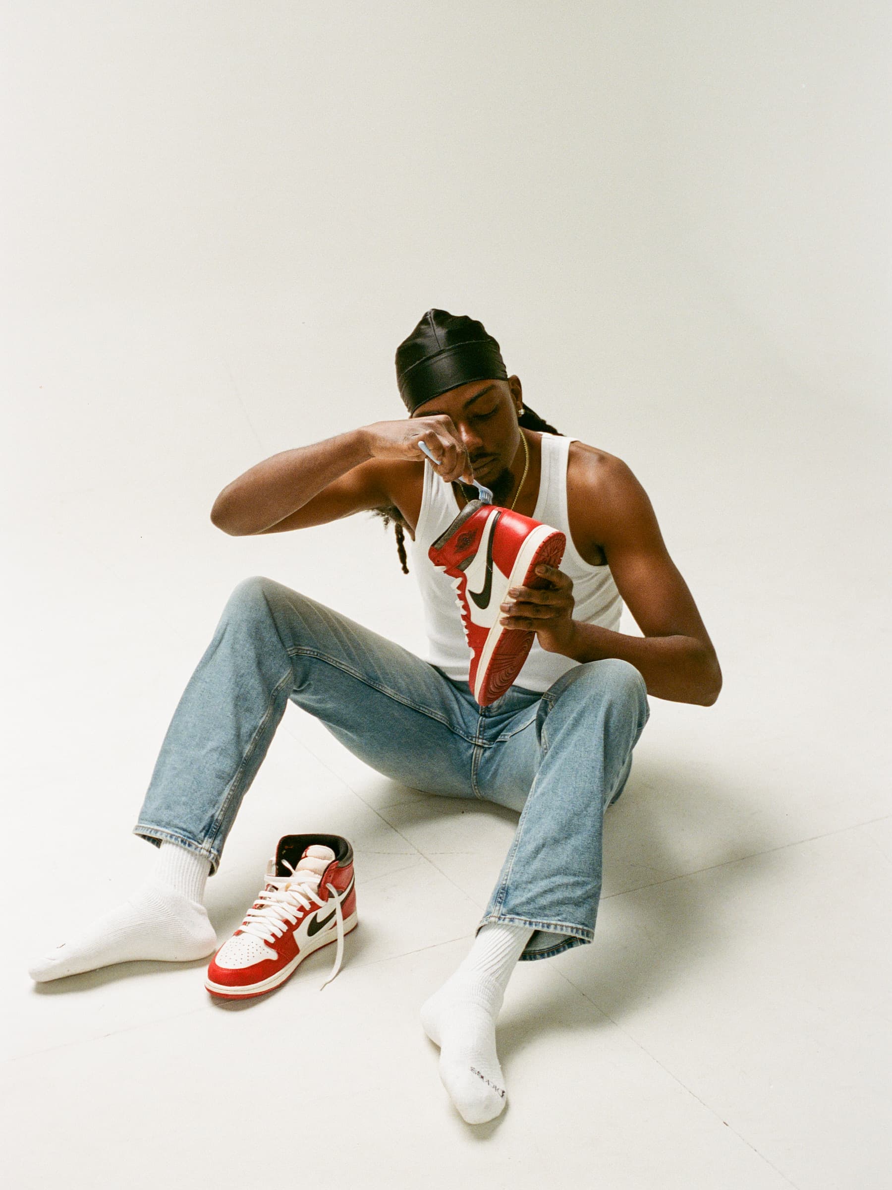 Portrait of a man in a black durag and white undershirt, cleaning a pair of sneakers with a toothbrush