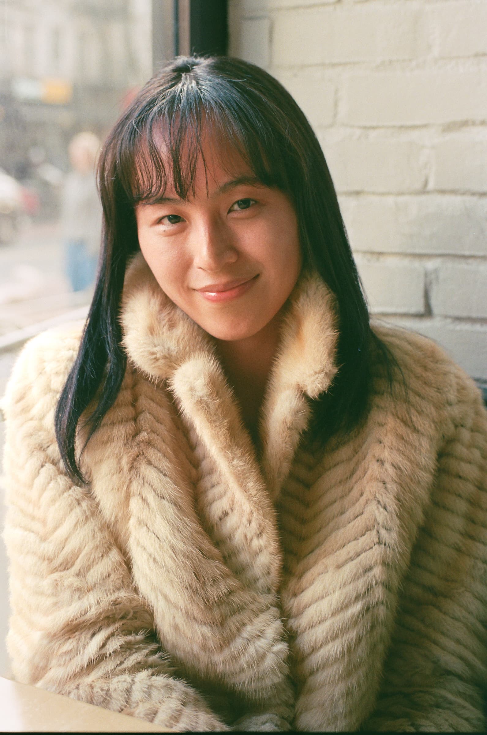 Portrait of a woman with long black hair and a fur jacket, seated inside a restaurant