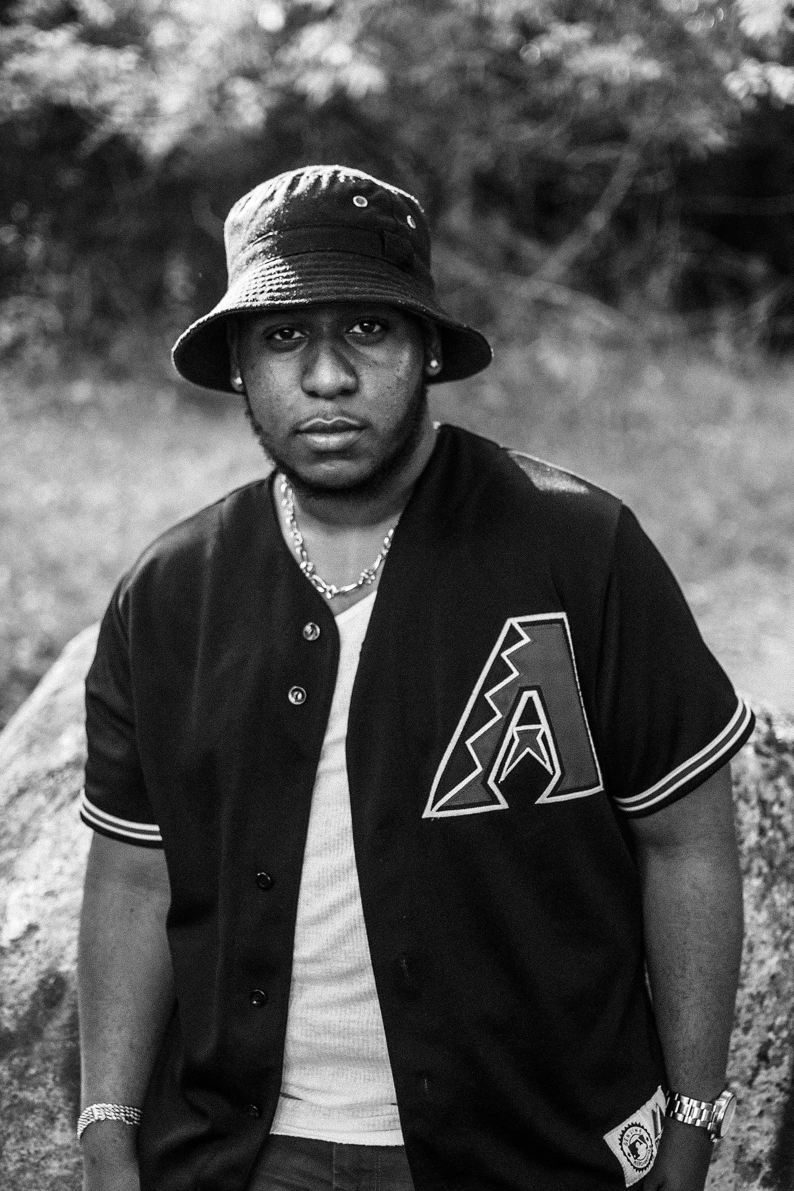 Black and white portrait of a man in a bucket hat and baseball jersey, standing outdoors in front of a large rock