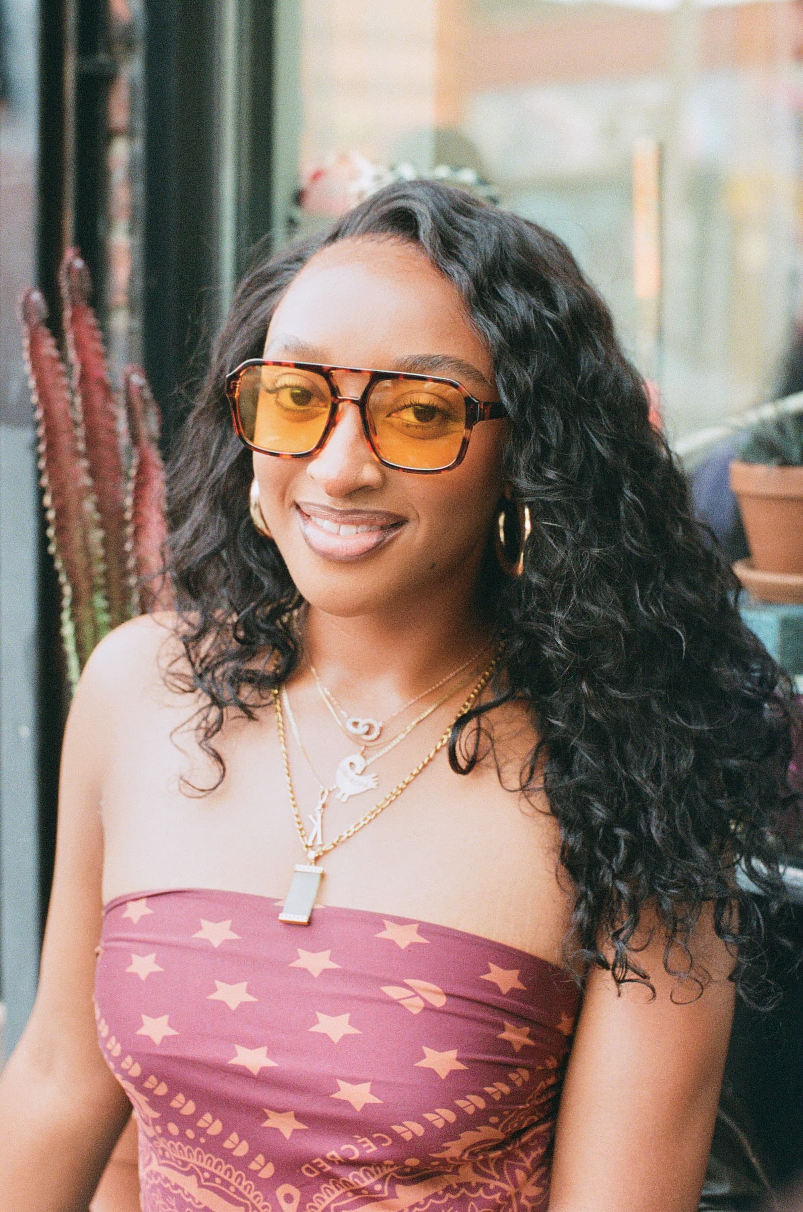 Portrait of a woman in a red top and orange glasses, seated in front of a storefront, smiling